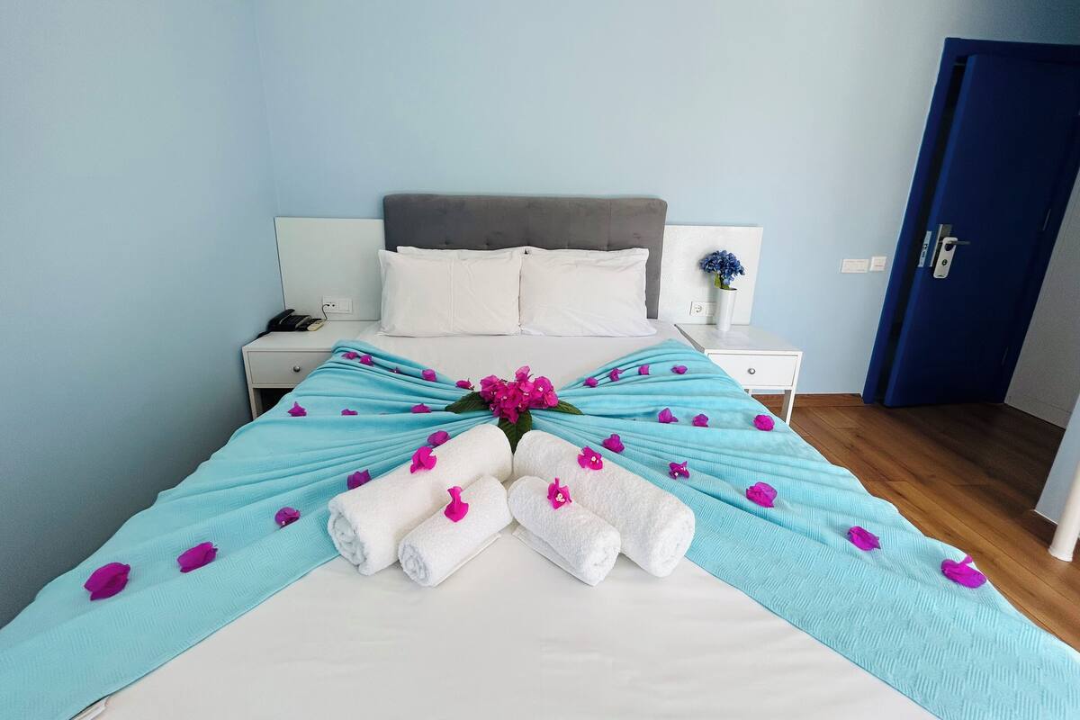 Een net opgemaakt bed met een blauw-witte dekbedovertrek, witte kussens en een boeket roze bloemen.