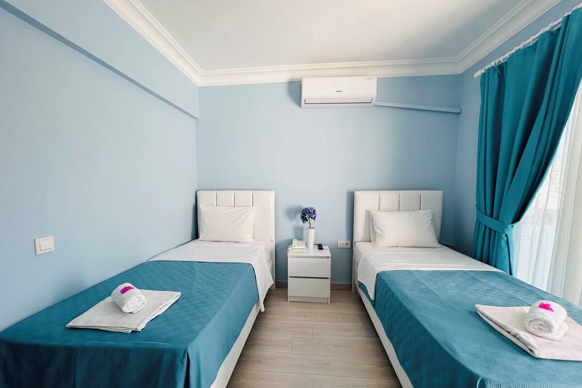 Een hotelkamer met twee bedden, een nachtkastje, een airconditioning en turquoise gordijnen.