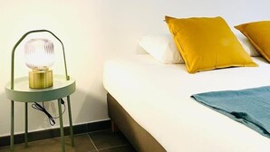 Una cama bien hecha con almohadas blancas y mostaza, una manta azul y una lámpara de noche sobre una mesita.