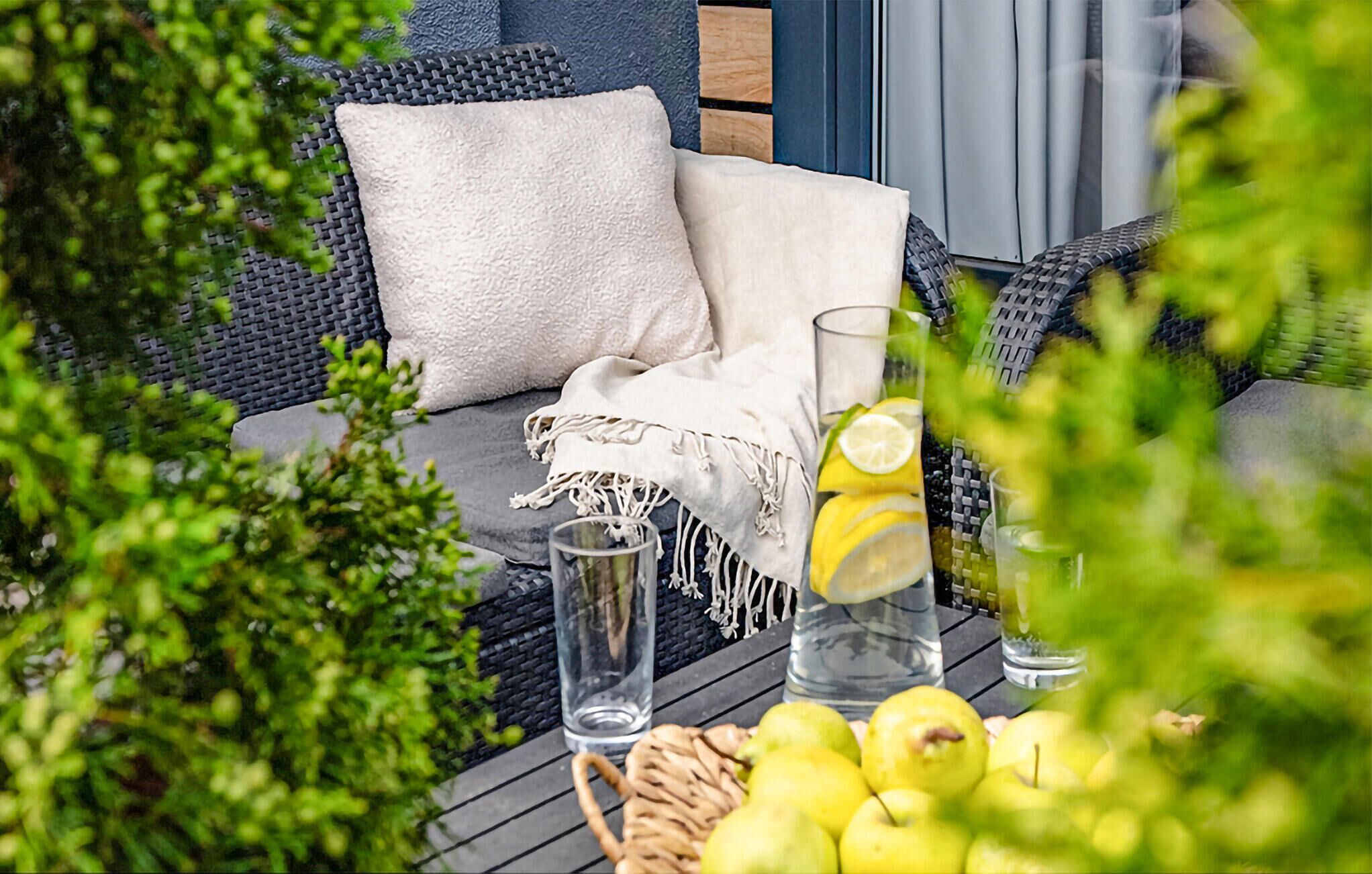 Un canapé en osier avec des coussins, un verre d’eau citronnée et un panier de pommes sur une terrasse en bois.