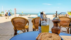 A table with a bottle of Blanche de Namur beer, a wooden salt shaker, and slices of bread on a seaside terrace with wicker chairs and a view of the beach. - Lovely apartment in Oostende (Oostende)