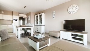 A modern living room with a flat-screen TV, a dining area with a white table and chairs, a kitchen with white cabinets, and a large wall clock. - Lovely apartment in Oostende (Oostende)