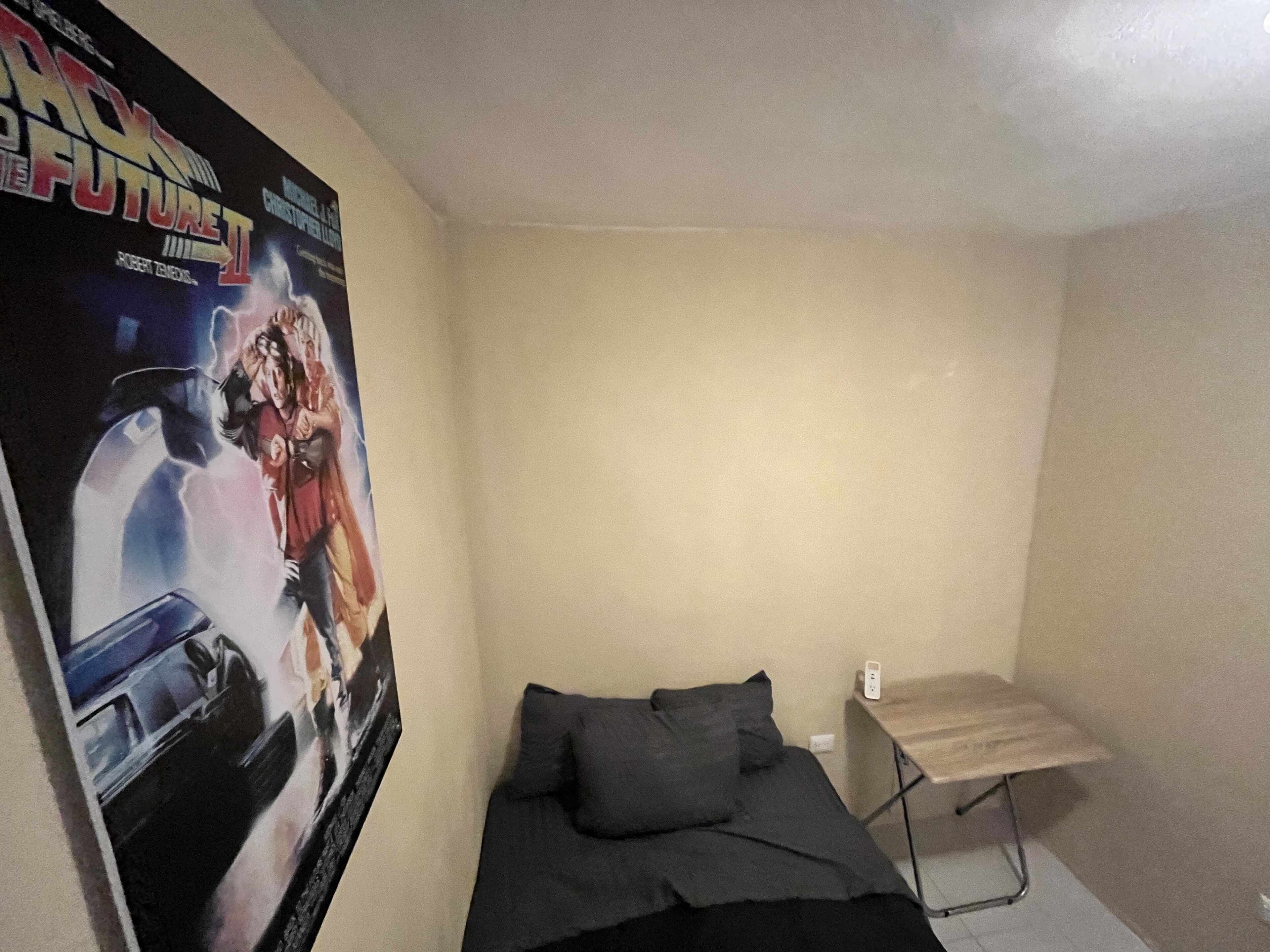 Una habitación pequeña y escasamente amoblada con una cama individual, una mesita y un cartel en la pared.