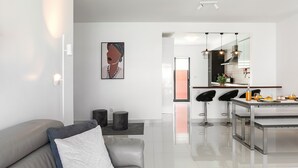 A modern living room with a grey sofa, a white cushion, and a coffee table. There is a kitchen area with a dining table, bar stools, and a large mirror above the counter. - Baleal Deluxe Flat - Shared pool (Peniche)