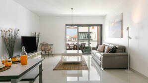 A modern living room with a sofa, coffee table, and a TV on a stand. There is a dining area with chairs and a table, and a view of the outside through a glass door. - Baleal Deluxe Flat - Shared pool (Peniche)