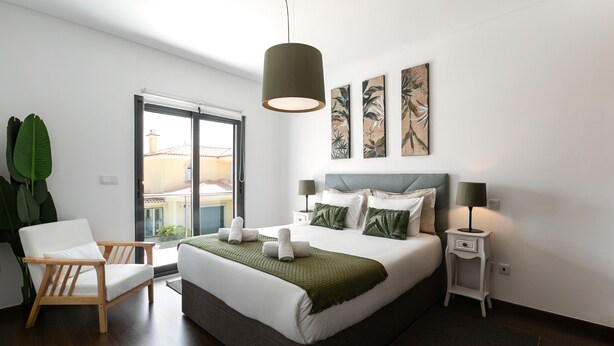 A modern bedroom with a large bed, a wooden chair, a bedside table with a lamp, and artwork on the wall. - Baleal Deluxe Flat - Shared pool (Peniche)