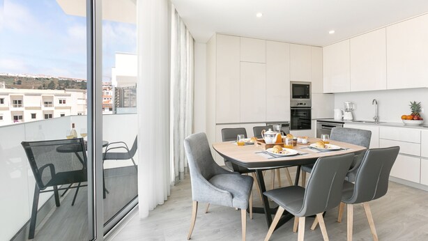 Een moderne keuken met witte kasten, een eethoek met een ronde tafel, en een uitzicht op gebouwen door grote ramen.