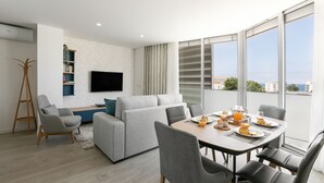 Een moderne woonkamer met een grijze bank, een eettafel met stoelen, een aan de muur bevestigde televisie, en een groot raam met uitzicht op gebouwen en de zee.