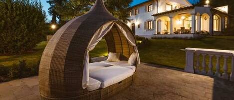 Un espacio exterior acogedor para dormir con un dosel tejido, una cama cómoda y una casa bien iluminada al fondo.