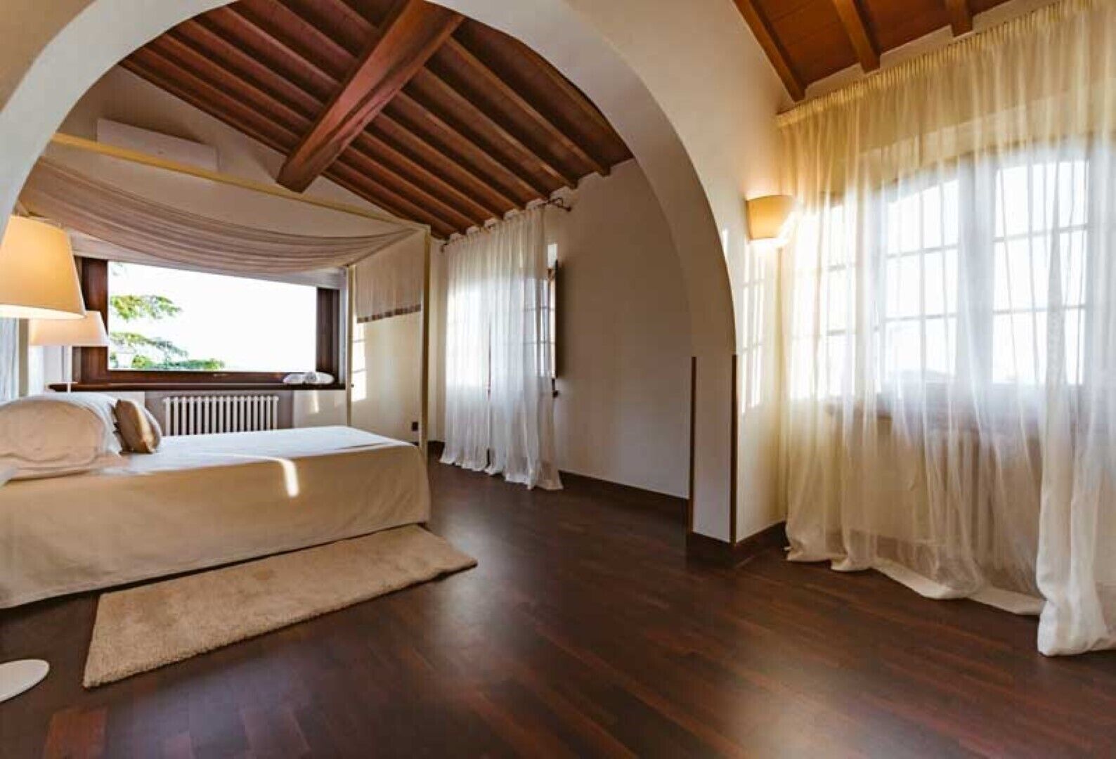 A bedroom with a large bed, wooden beams, and a view of greenery outside.