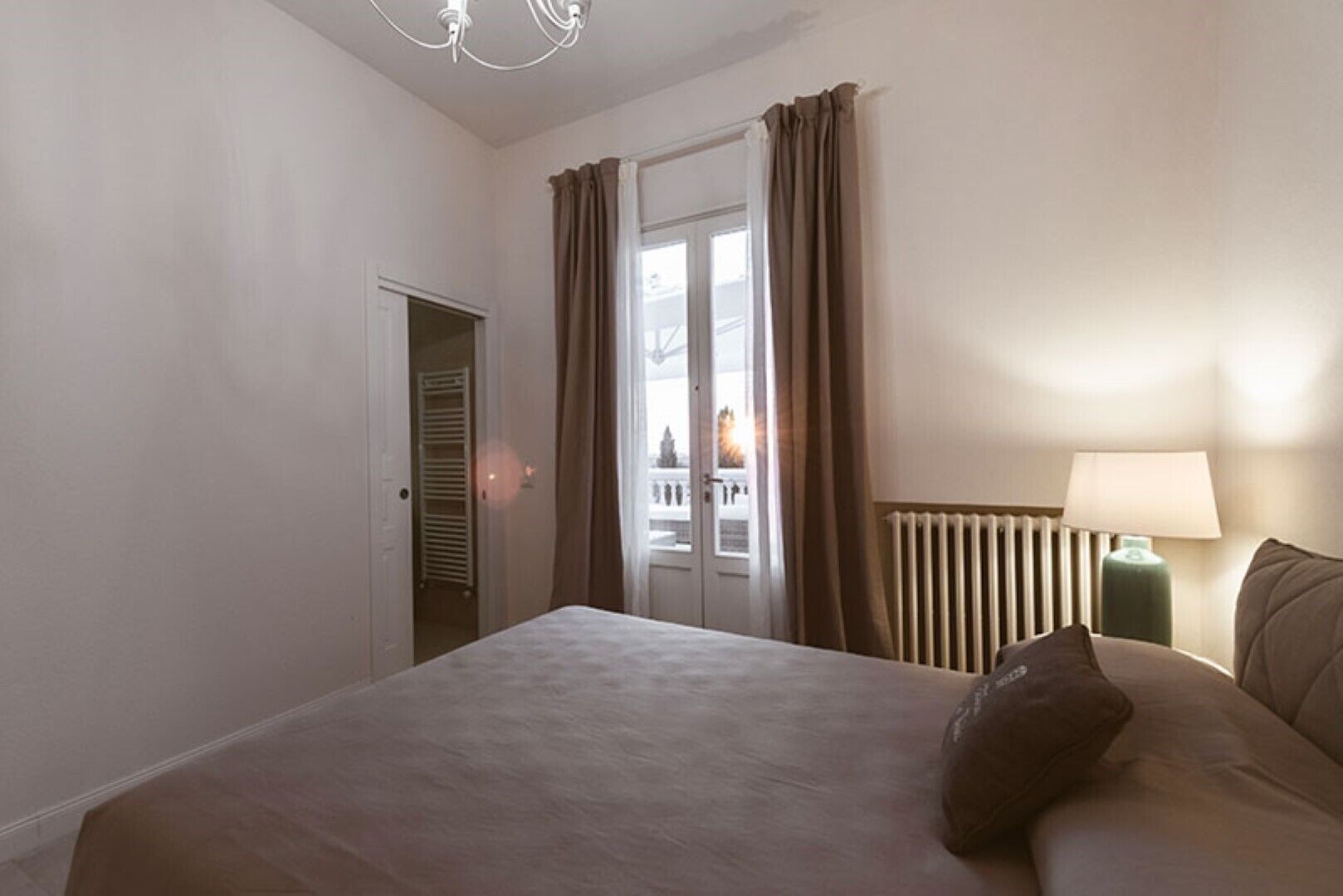 A bedroom with a bed, a nightstand with a lamp, a radiator, and a view through a door.