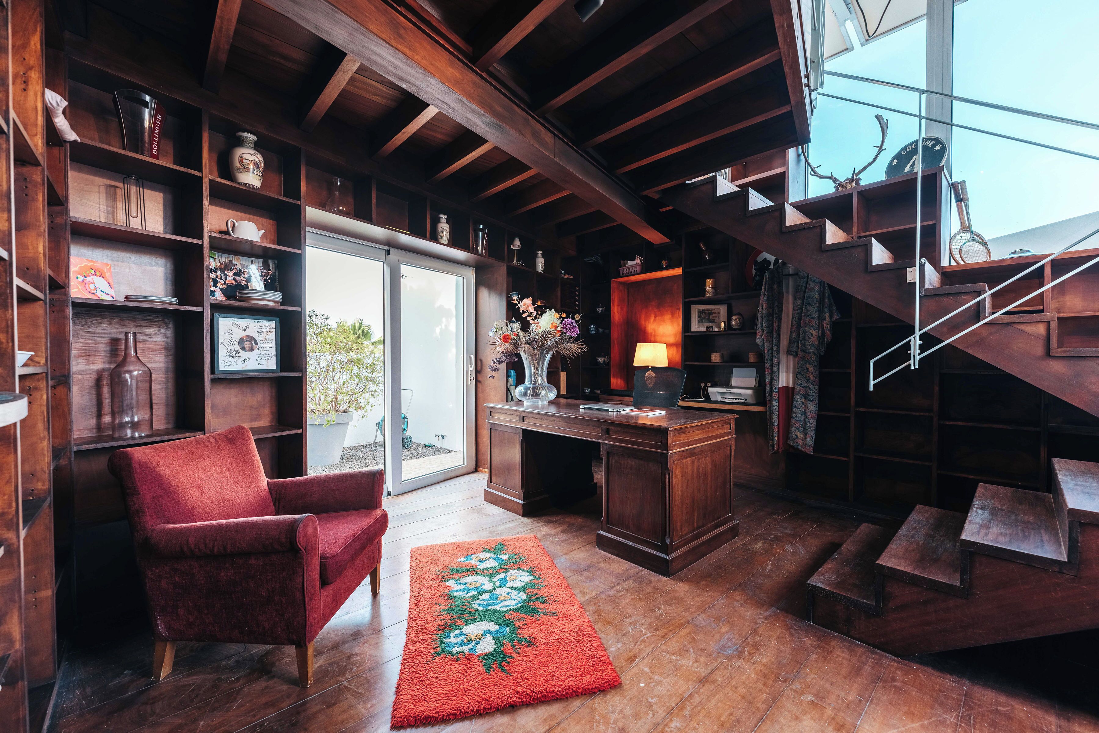 A cozy room with a wooden desk, a red armchair, and a staircase leading to an upper level.