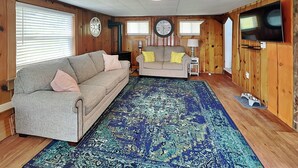 Una sala de estar acogedora con un sofá gris, una alfombra estampada, paredes de madera, un televisor y un reloj.