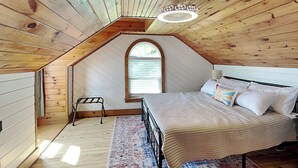 Un dormitorio con techo inclinado, una cama, una mesita de noche, una lámpara y un ventanal con persianas.
