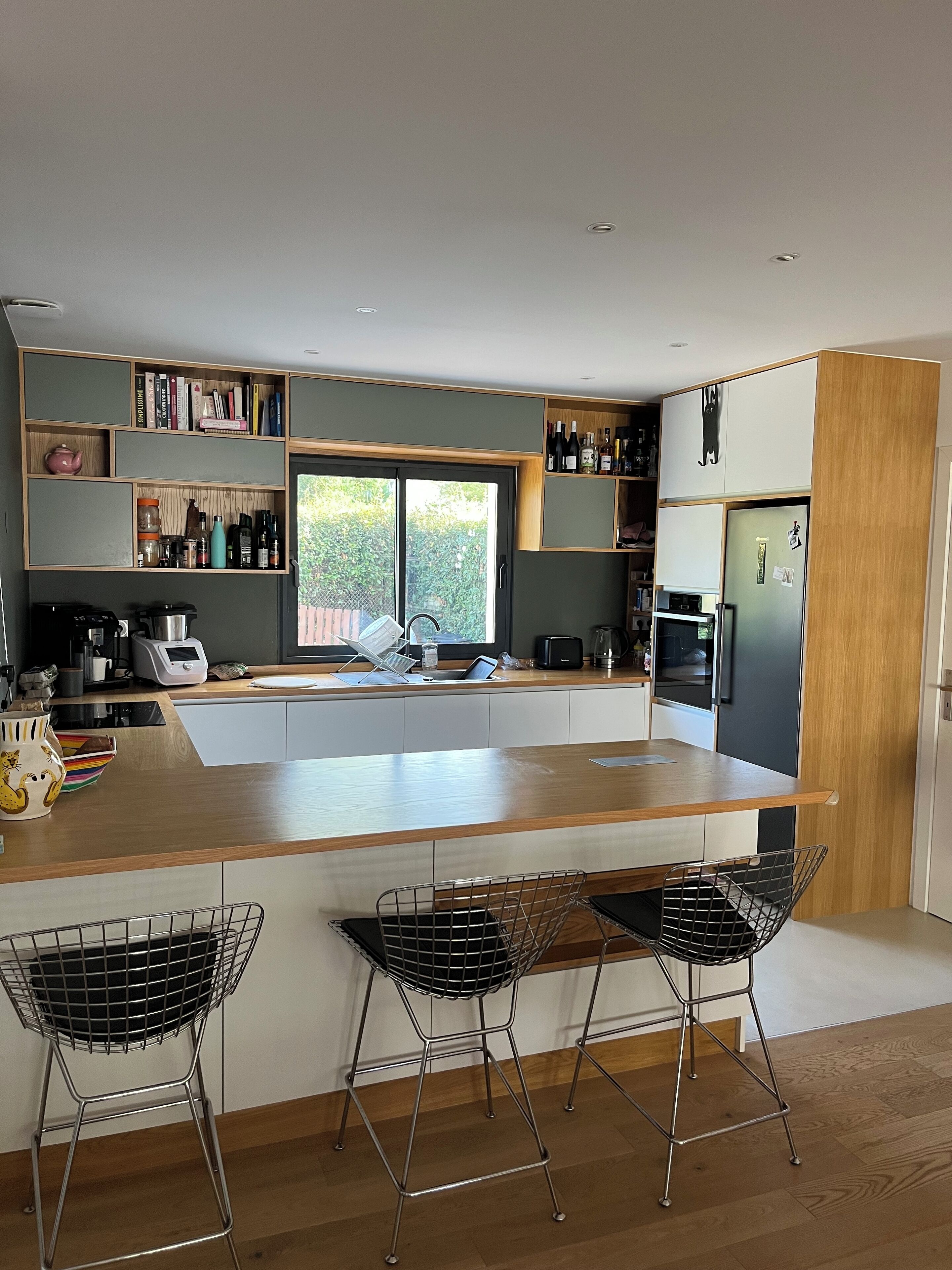 Een moderne keuken met een centraal eiland, barkrukken, inbouwplanken en een groot raam.