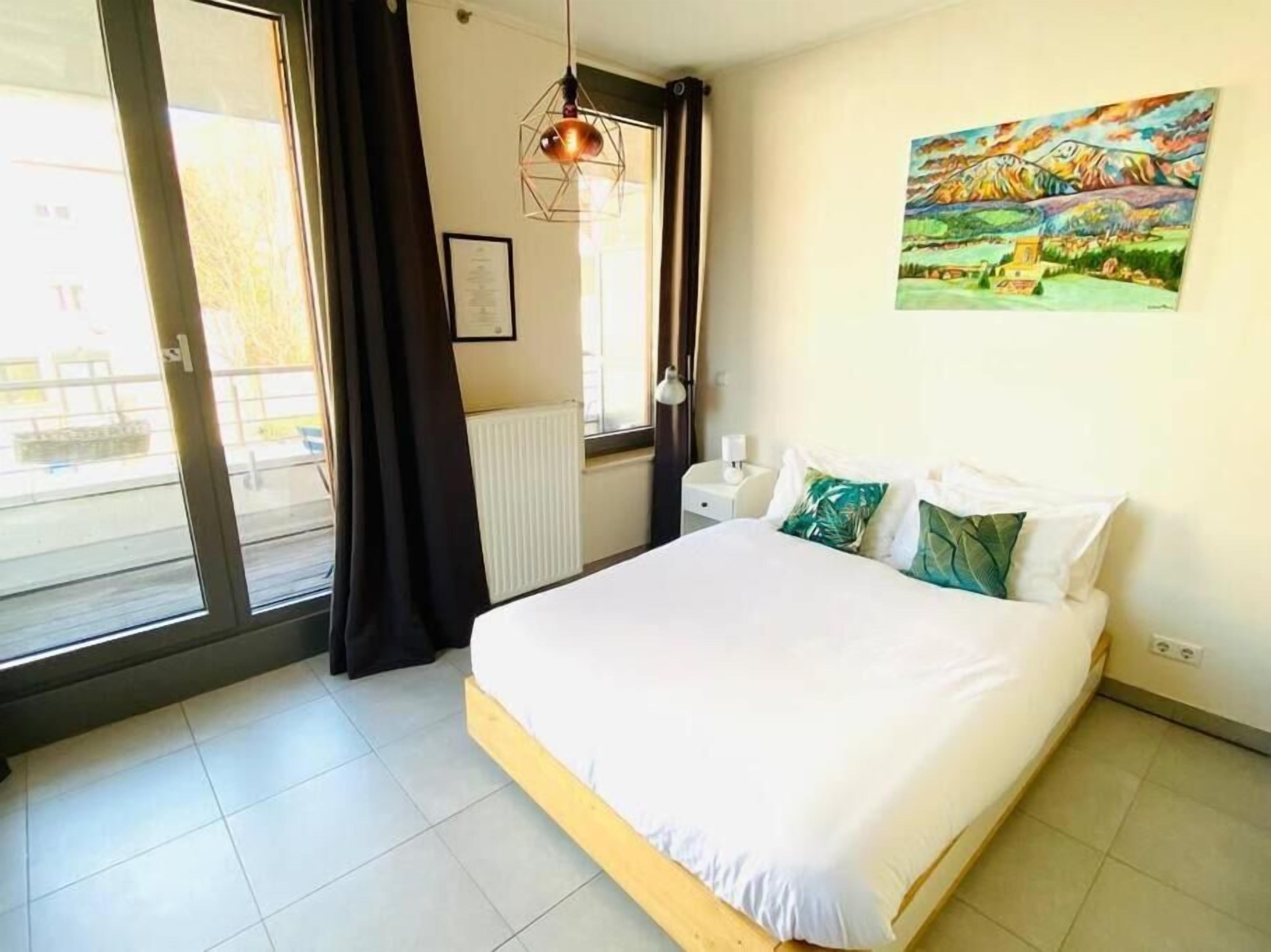 A bedroom with a large bed, a window with curtains, a radiator, a nightstand, and a painting on the wall.