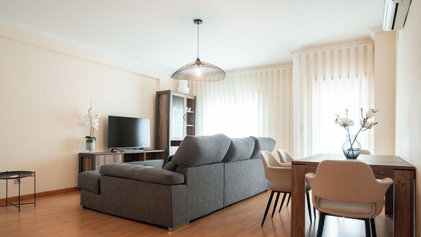 Una sala de estar moderna con un sofá gris, un televisor, una mesa de comedor con sillas y una lámpara colgante.