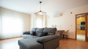 A modern living room with a grey sectional sofa, a dining table, and a kitchen area visible through an open door.