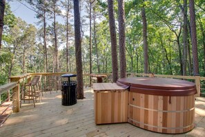 A wooden deck with a hot tub, chairs, and a trash can in a forest setting.