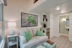 A living room with a light-colored sofa, green cushions, a side table with a lamp, a framed artwork, and a kitchen area with a microwave and cabinets.