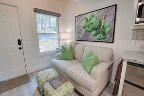 A living room with a beige sofa, green cushions, a patterned ottoman, a framed artwork of clover leaves, and a kitchen area with a granite countertop and stainless steel appliances.