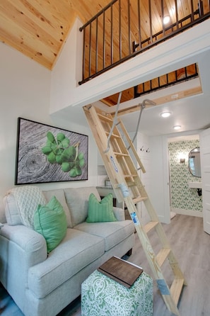 A loft-style living room with a wooden staircase, a light-colored sofa with green cushions, a framed artwork on the wall, and a small table with a book.