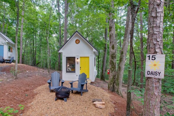 A small cabin with a yellow door, surrounded by trees, and two Adirondack chairs in front.