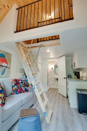 Ein Loft-Wohnraum mit einer Holztreppe zu einer erhöhten Ebene, einem Küchenbereich und einer Couch mit farbenfrohen Kissen.