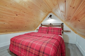 A bedroom with a sloped ceiling, wooden walls, a bed with red bedding, and a small window.