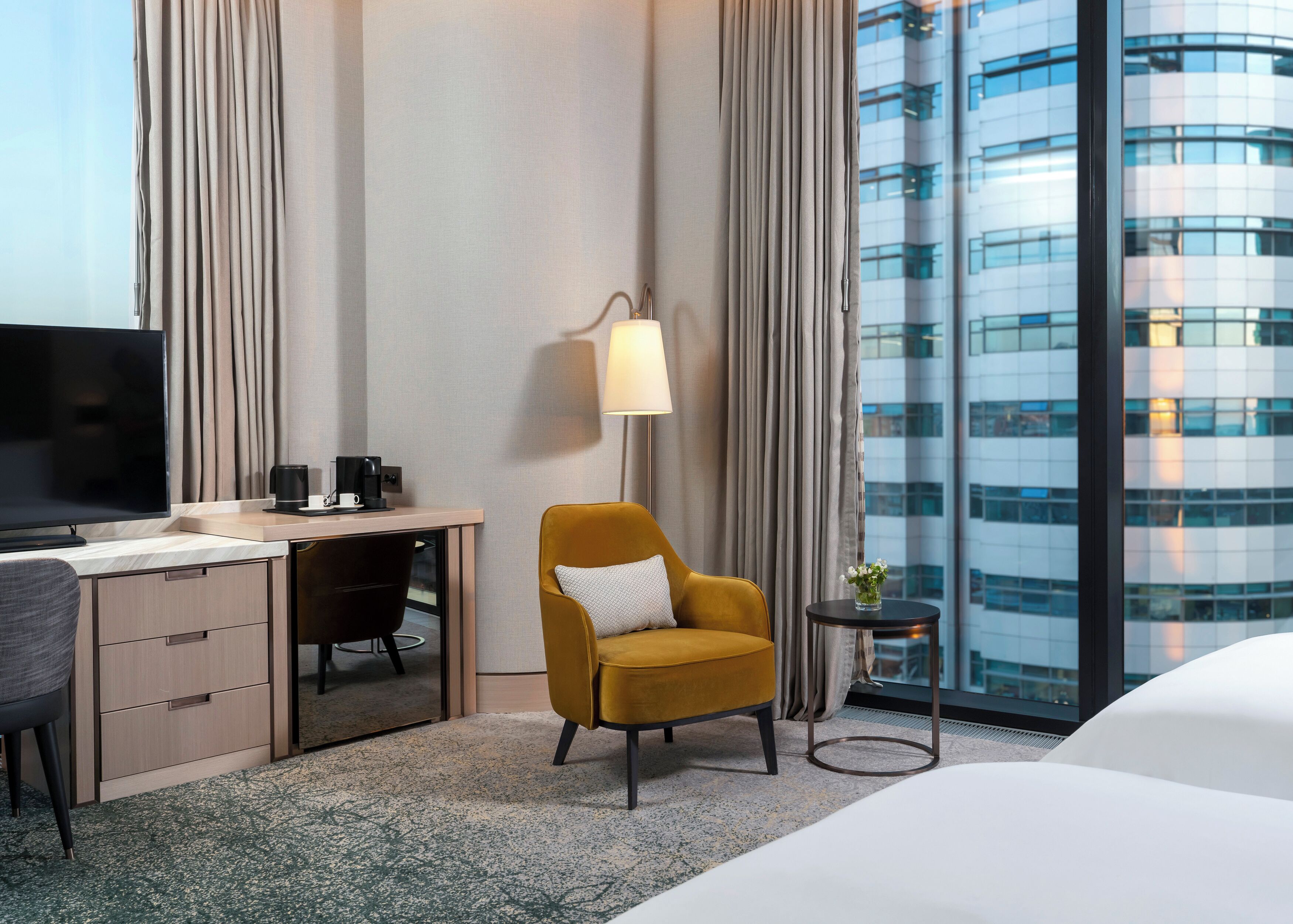 A hotel room with a bed, a desk with a TV, a chair, a lamp, and a view of a modern building.