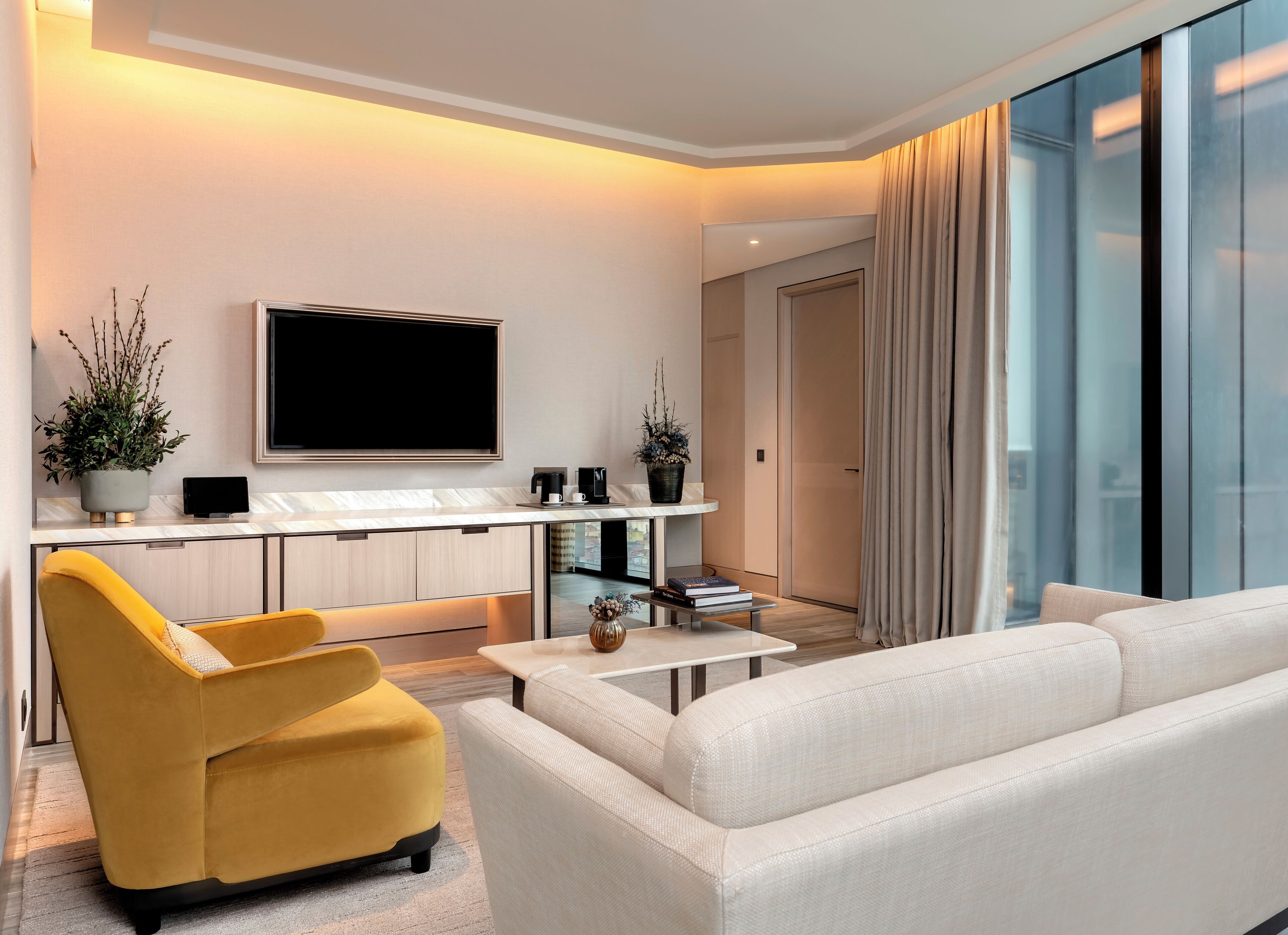 A modern living room with a sofa, a yellow armchair, a coffee table, a flat-screen TV, and a kitchen area with a countertop and cabinets.