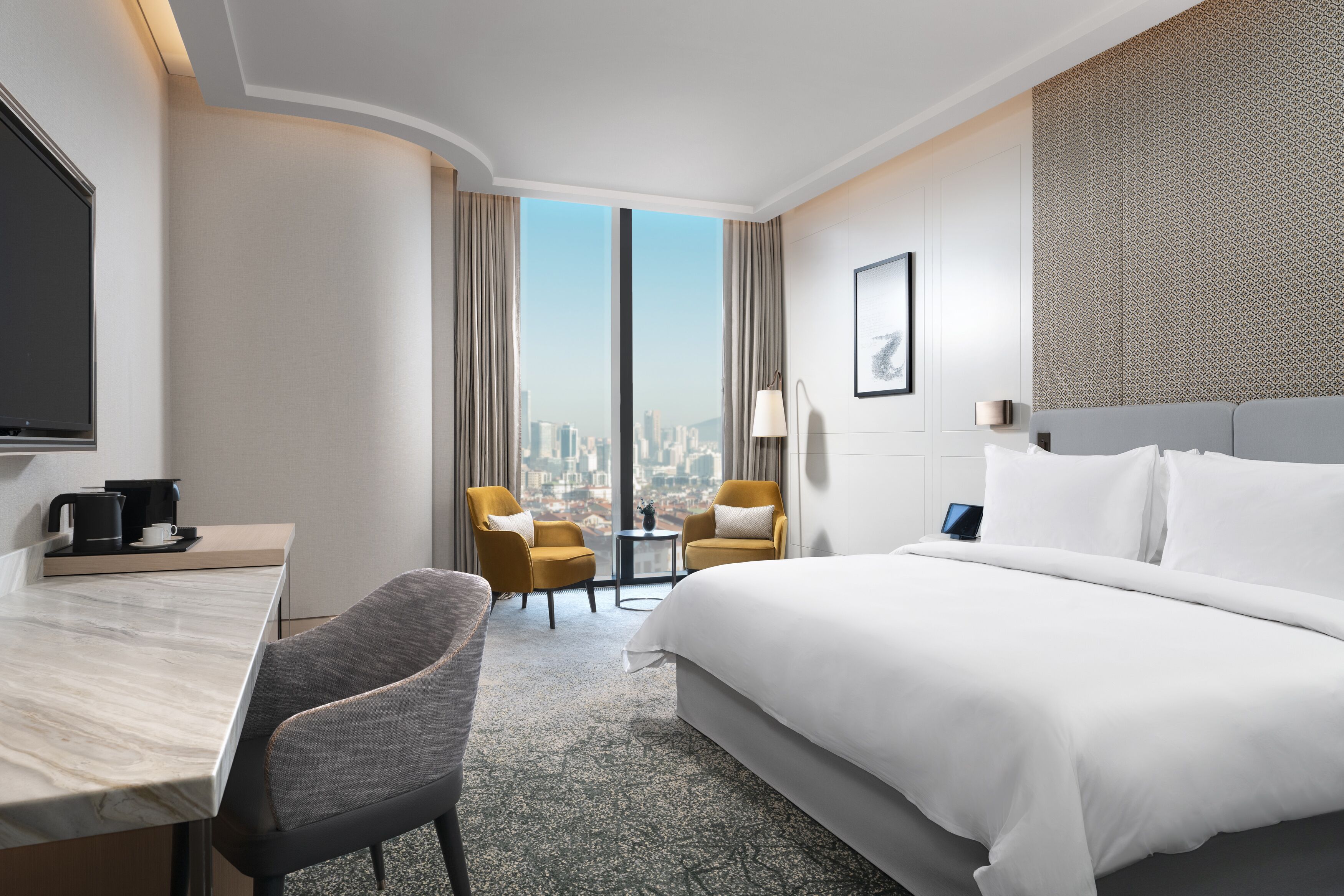 A hotel room with a large bed, a desk, two armchairs, a small table with a lamp, and a city view through large windows.