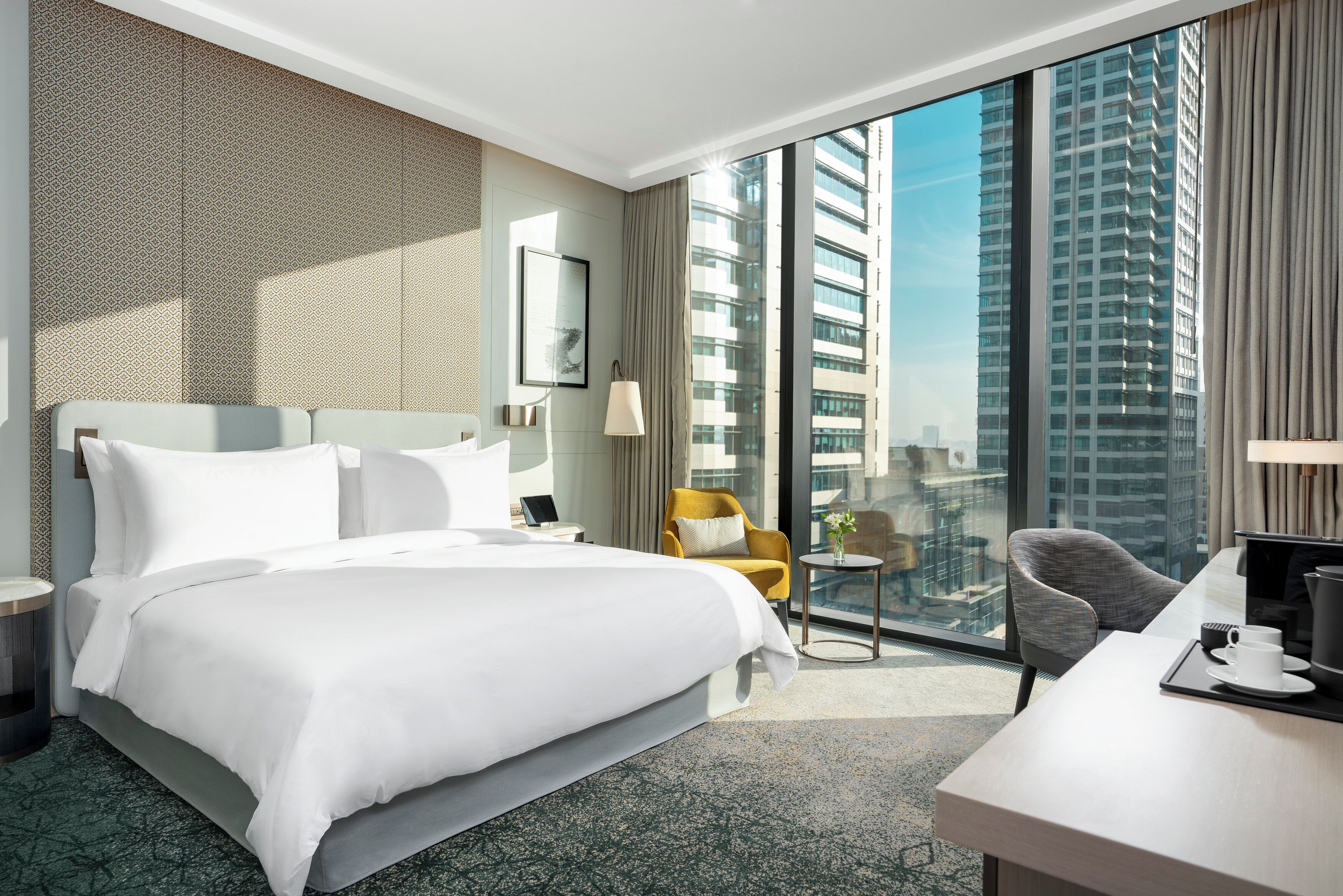 A hotel room with a large bed, a desk, a chair, and a view of tall buildings.