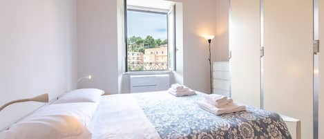 A bedroom with a bed, a window with a view of buildings, a bedside lamp, and a wardrobe.