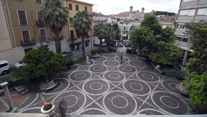 A courtyard with patterned paving, palm trees, and surrounding buildings. - Domo Peonia Bianca (Nuoro)