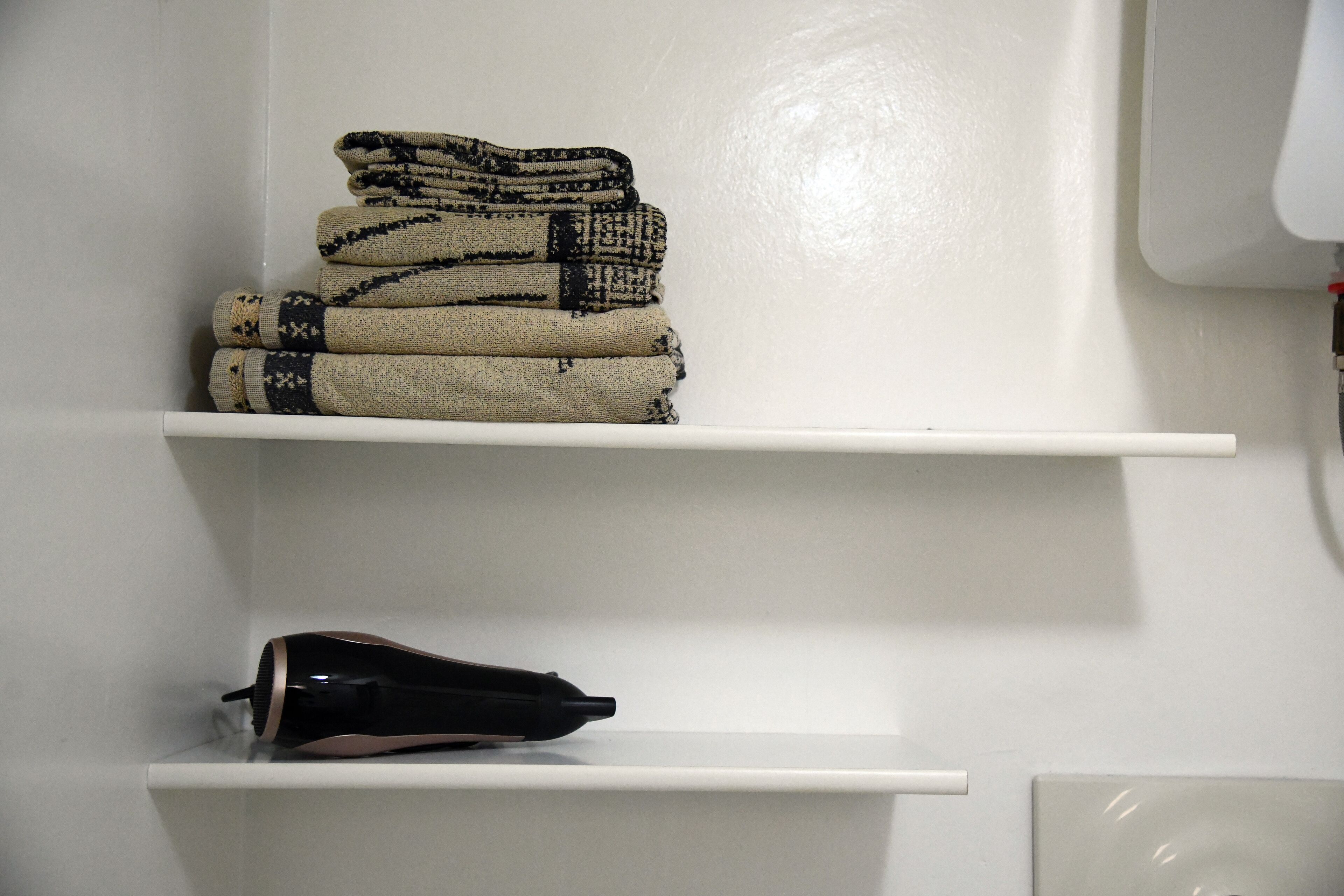 A white wall with two white shelves; one shelf holds folded blankets, and the other shelf holds a black object.