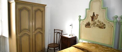 A bedroom with a bed, a wooden wardrobe, a chair, and a nightstand with a lamp.
