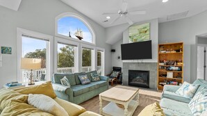 A spacious living room with a fireplace, large windows, and a ceiling fan. - 1528 - Sound and Color - Spacious Soundfront Retreat w/ Stunning Sunsets (Corolla)