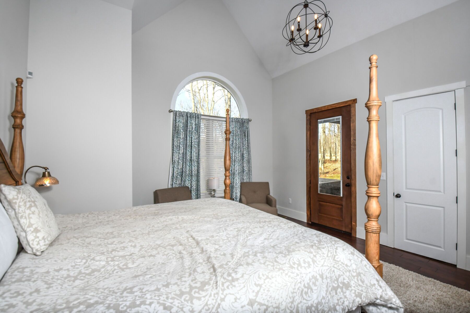 A bedroom with a large bed, a wooden four-poster, a chandelier, a door with a view, and a window with curtains.