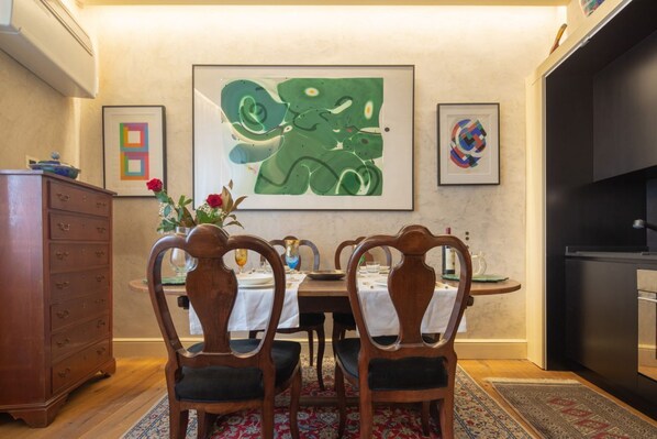 Uma sala de jantar com uma mesa posta para quatro pessoas, cadeiras de madeira, um vaso com flores e arte abstrata nas paredes.