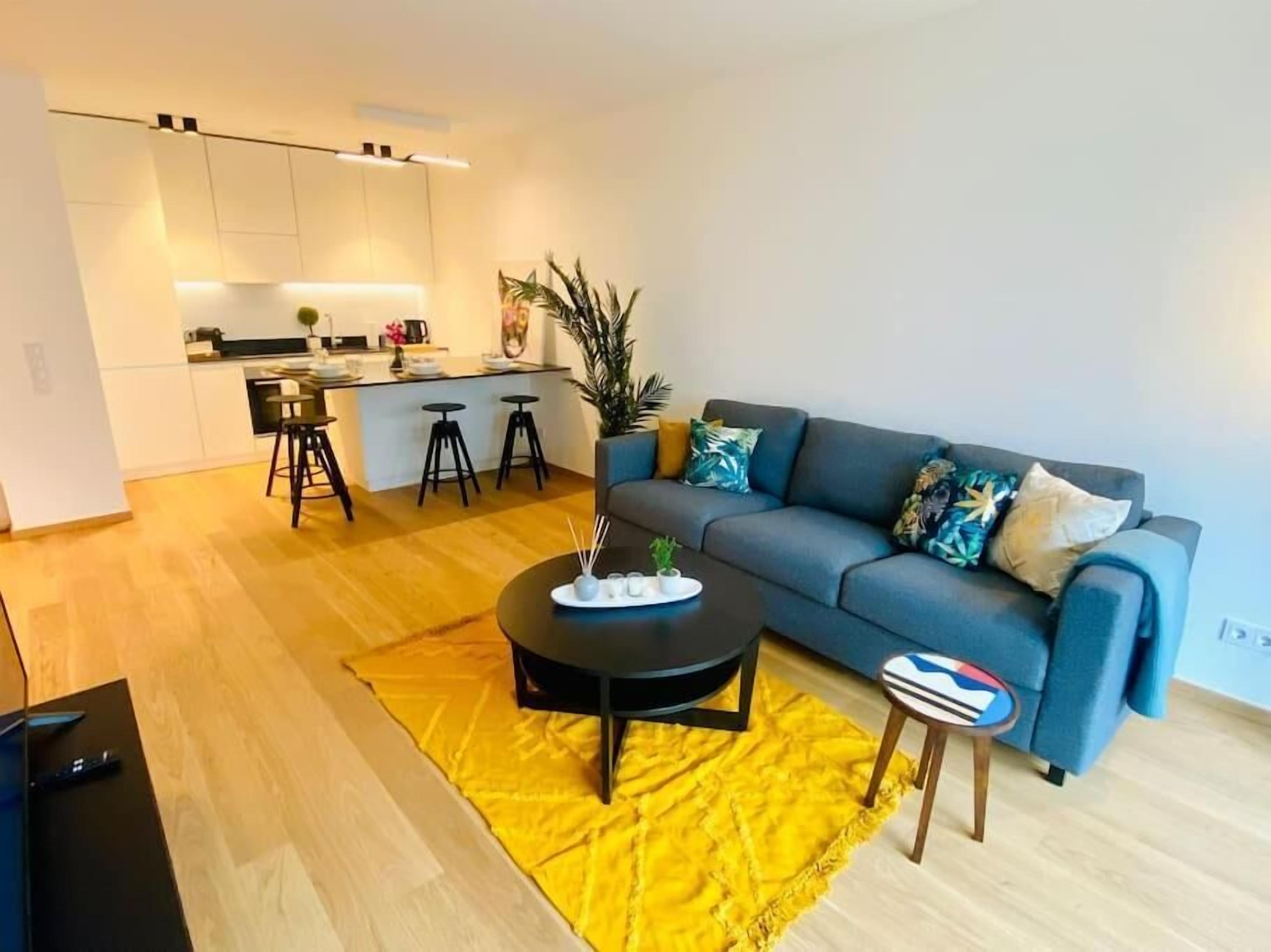 Una sala moderna con un sofá, una mesa de centro y una mesita auxiliar. Se observa una zona de cocina al fondo con un taburete y una planta en maceta.
