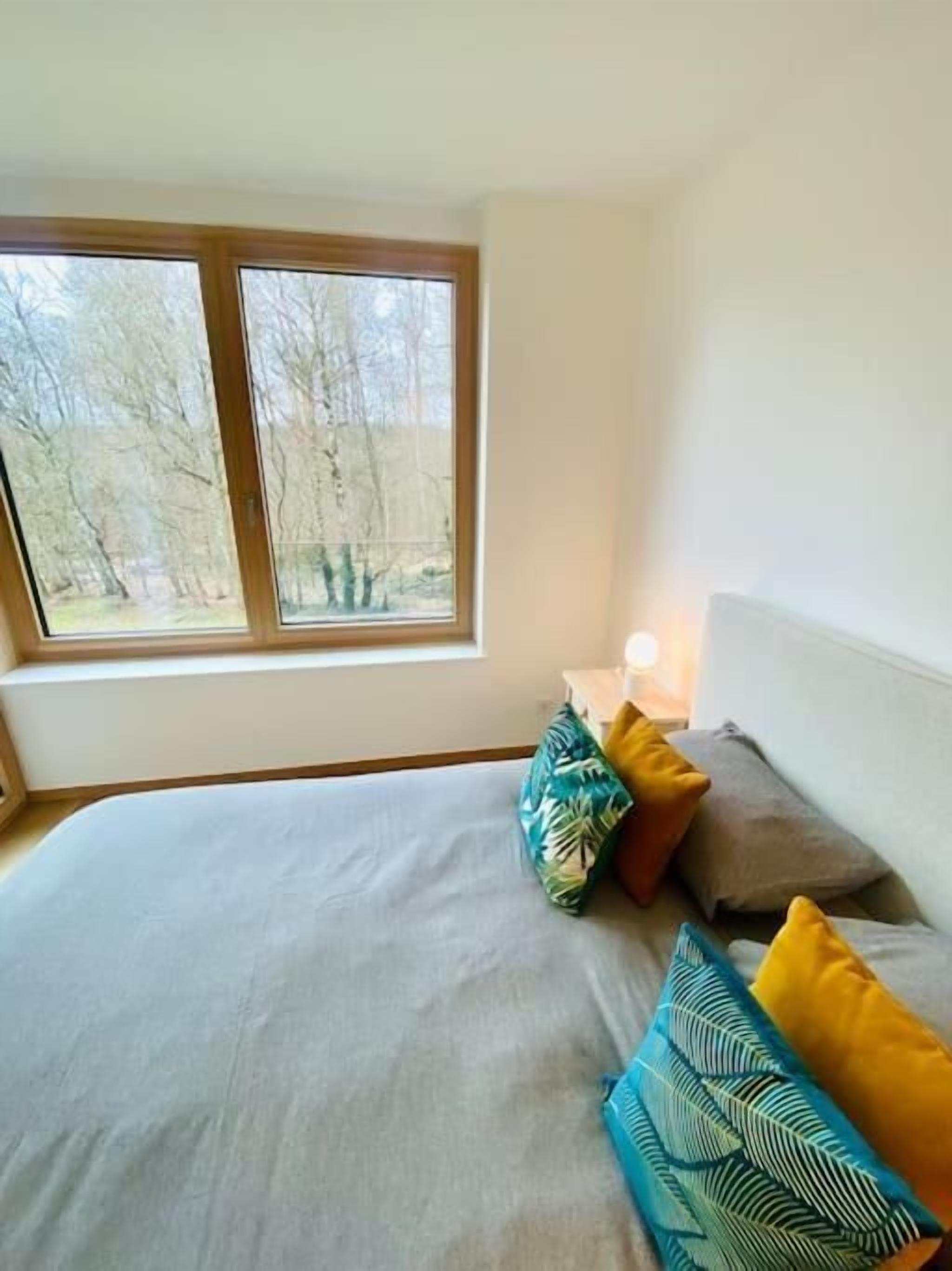 Een slaapkamer met een groot bed, meerdere kussens, een nachtkastje met een lamp en een raam met uitzicht op bomen.