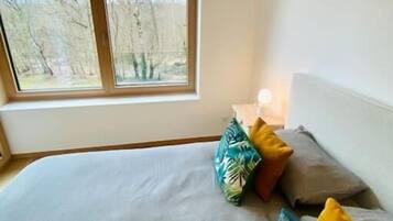 Een slaapkamer met een groot bed, meerdere kussens, een nachtkastje met een lamp en een raam met uitzicht op bomen.