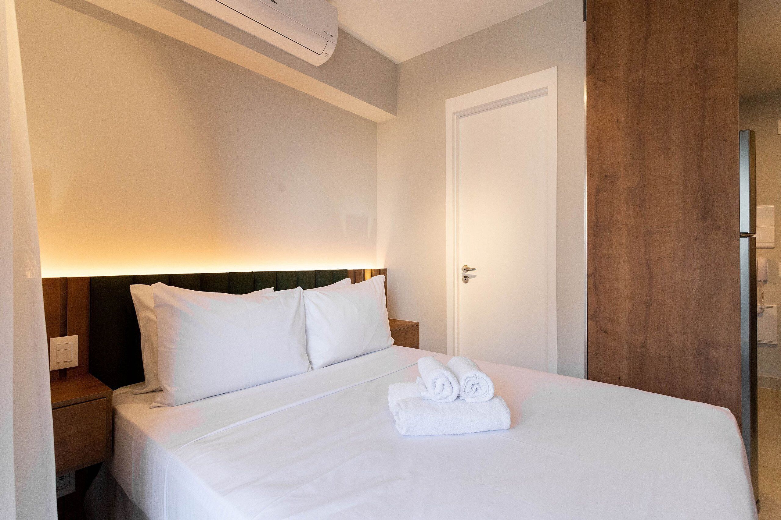 A neatly made bed with white linens and rolled towels, a wooden headboard, a bedside table with a lamp, and a door to the right.