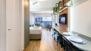 Ein modernes Hotelzimmer mit einem Bett, einem Esstisch mit Stühlen, einer Küchenzeile und einem großen Fenster.