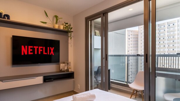 Ein modernes Hotelzimmer mit einem großen Flachbildfernseher, der "NETFLIX" zeigt, einem Bett mit ordentlich gefalteten weißen Handtüchern, einer Schiebetür zu einem Balkon mit einem Stuhl und einer kleinen Ablage mit Dekorationen.