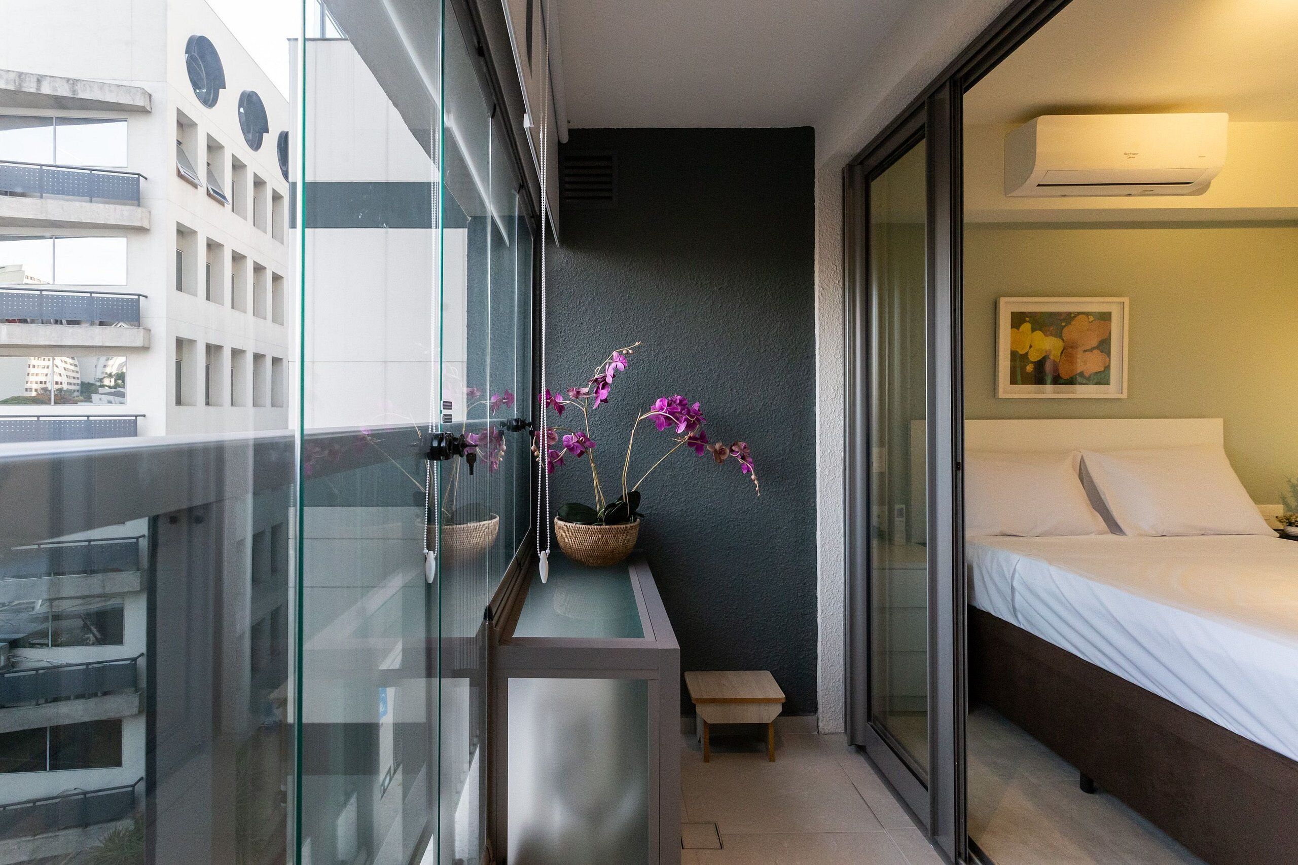 A modern hotel room with a balcony, a bed, a small bedside table, and a potted orchid plant.