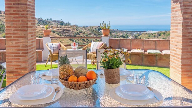 Un área de comedor al aire libre con una mesa preparada para dos personas, que incluye una cesta de frutas, una planta en maceta y vistas del paisaje.