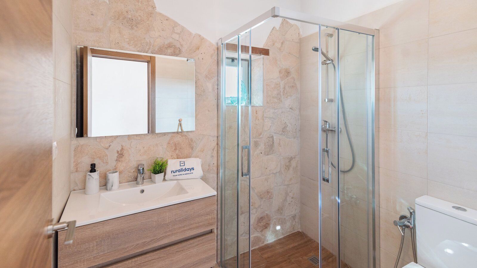 A modern bathroom with a stone wall, a glass shower enclosure, a wooden vanity, and a white sink.