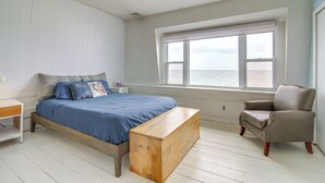 Un dormitorio con cama, un banco de madera, una silla, una mesita de noche y ventana con vista al mar.